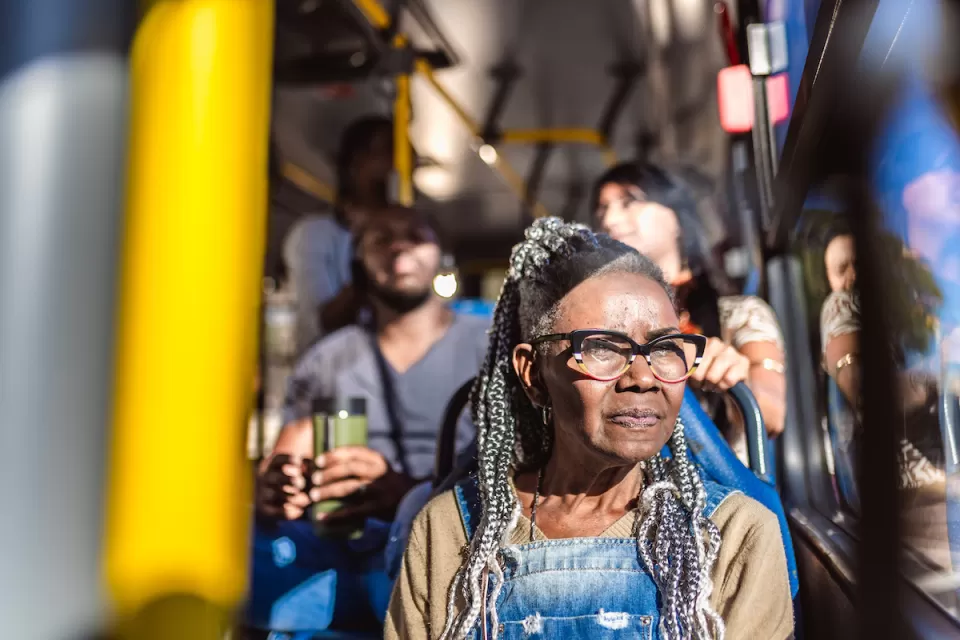 Older Black woman with glasses and braids sitting on city bus, looking out window.